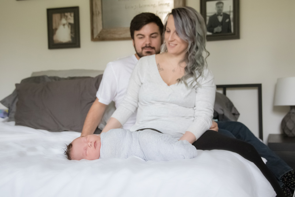 mom and dad sitting on bed looking at newborn baby
