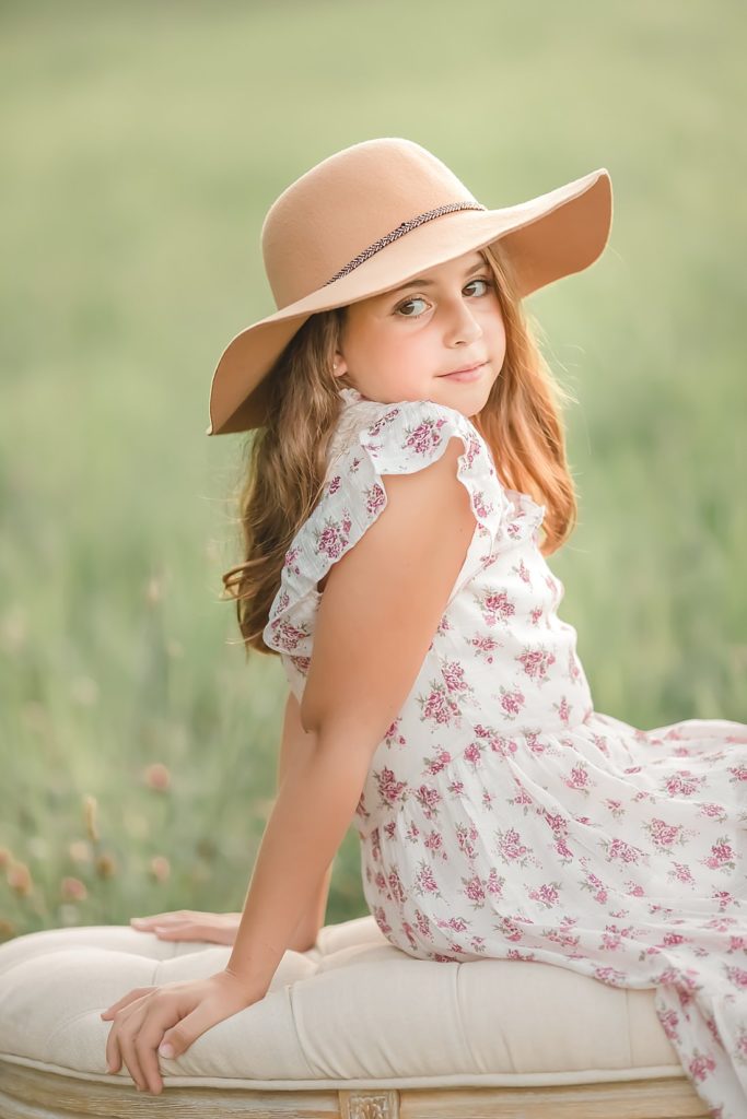 girl in floral dress and hat sitting on bench in field at sunset_petite magnolia photography