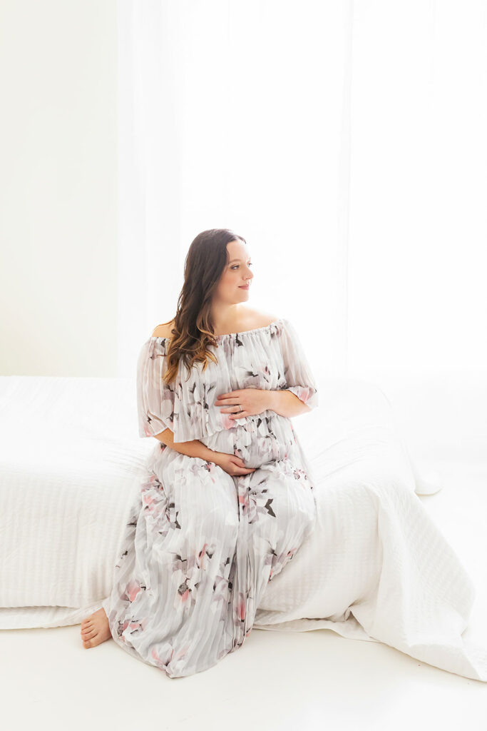 mother to be sits on a bed in a studio looking out the window