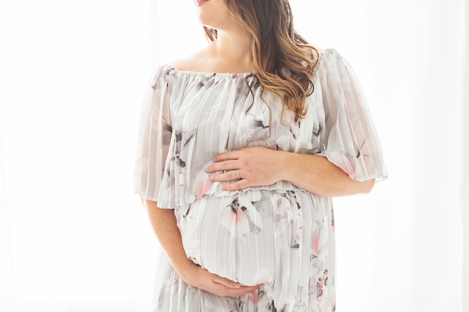 Mother to be in white flower print maternity gown stands in a studio holding her bump Pittsburgh Midwives