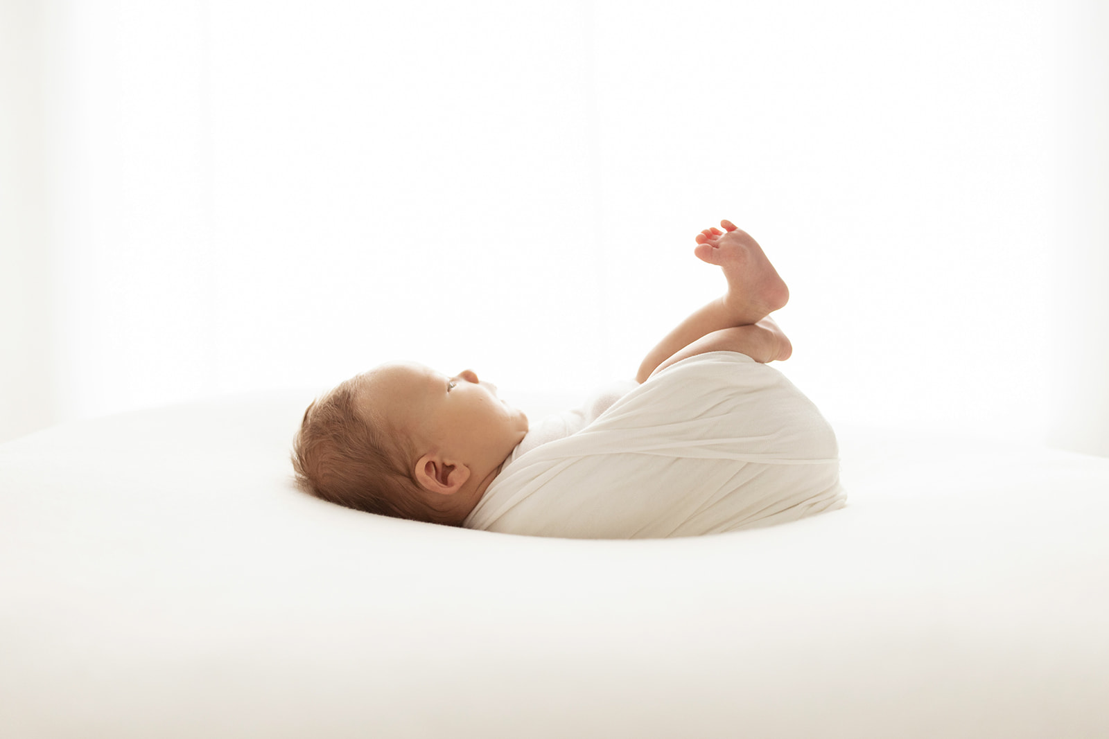 A newborn baby sleeps on it's back in a white swaddle with feet sticking up in the air