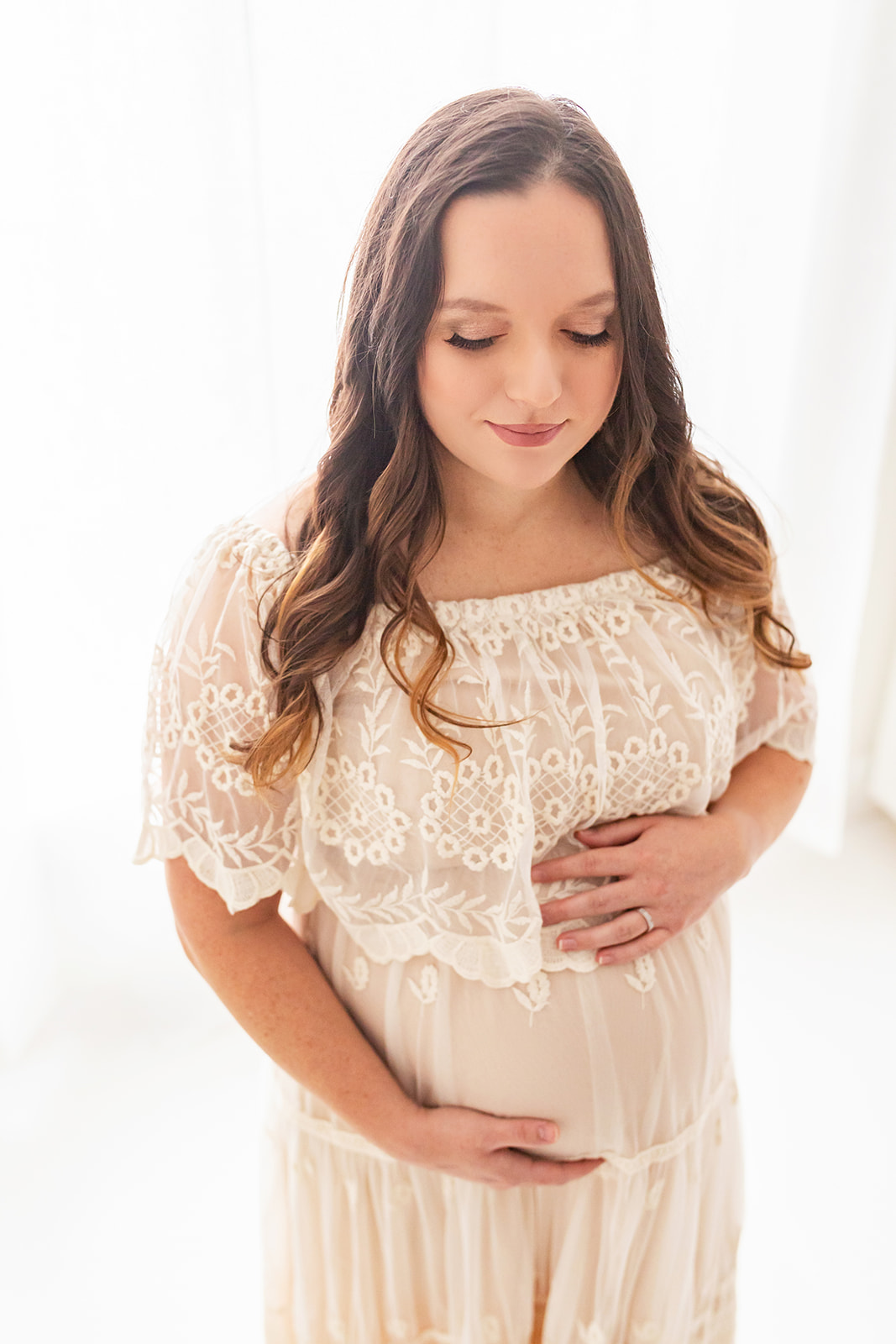 mother to be stands in a cream lace maternity gown looking down at her hands on her bump in front of a window midwife center pittsburgh