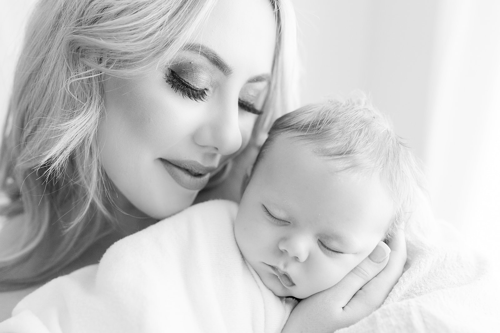 A mother with long blonde hair snuggles with her sleeping newborn baby