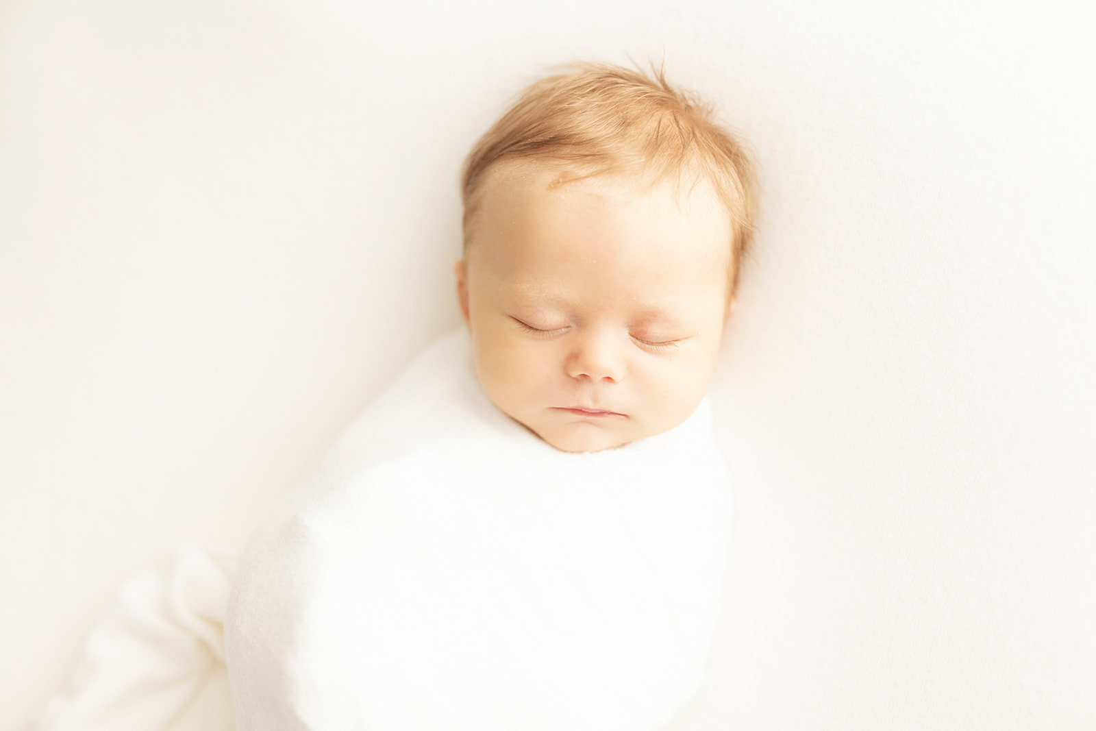 A newborn baby with blonde hair sleeps in a white swaddle next to a window