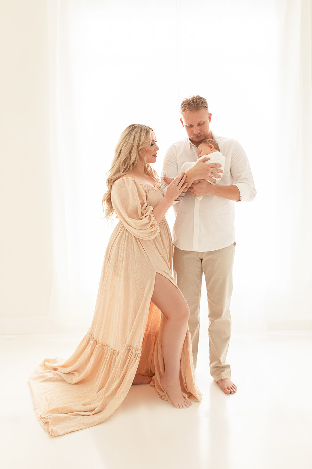 A father in a white shirt and khaki pants holds his sleeping newborn baby against his chest in front of a large window while mom in a flowing beige dress leans in to them Tullabee