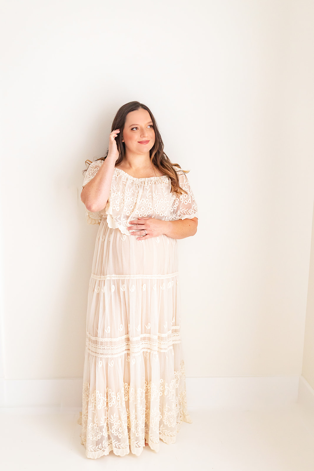 A mother to be stands in a studio while wearing a lace maternity gown