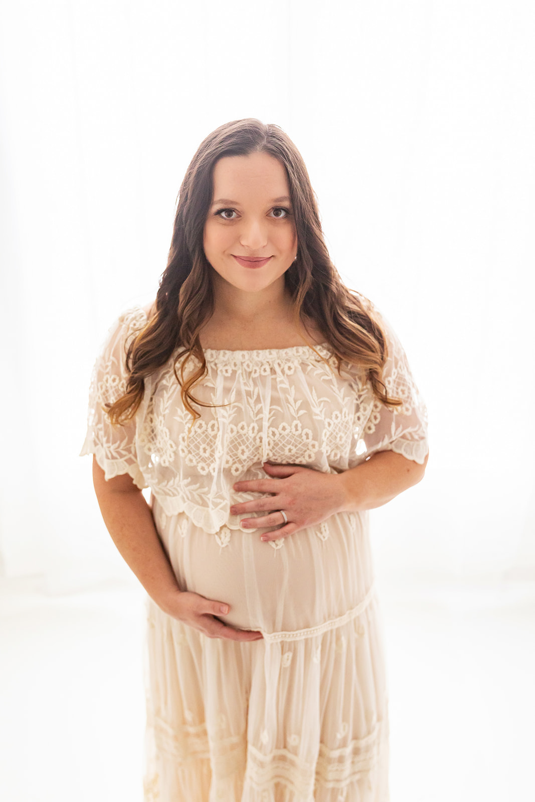 A mom to be holds her bump while standing in front of a window with long brown hair and a lace maternity dress The Steel Orchid