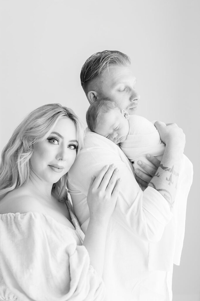 A father stands in a white shirt holding his sleeping newborn on his shoulder while mom leans on him All About Kids Furniture