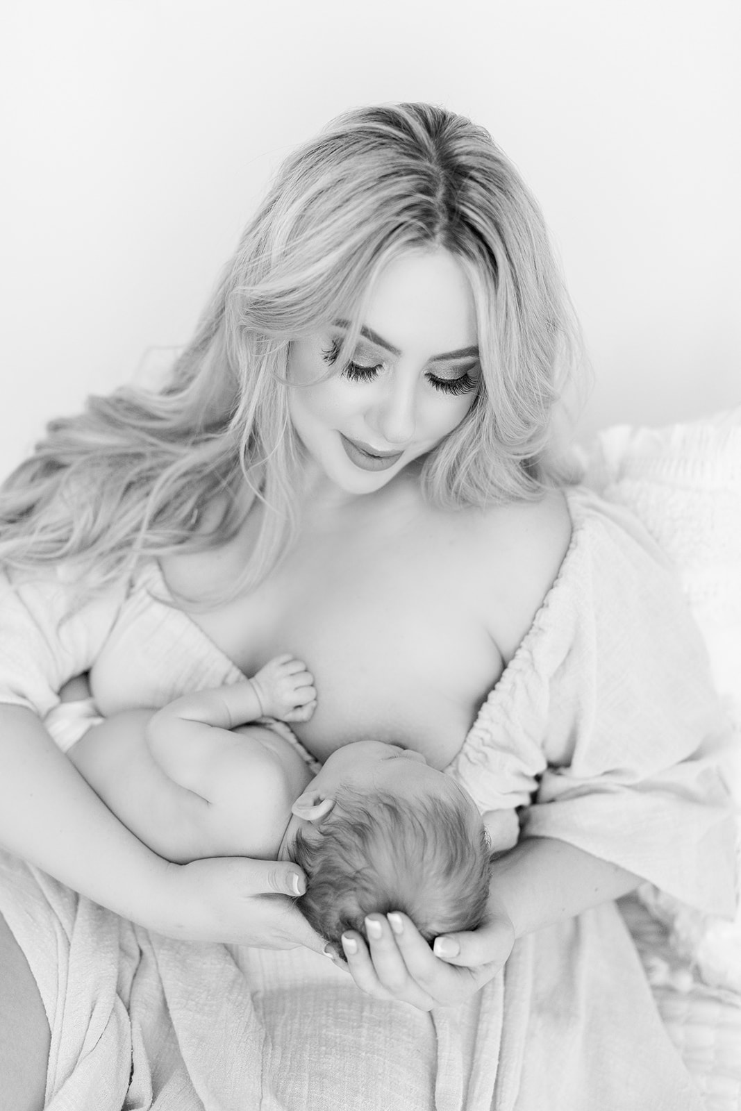 A mother breastfeeds her newborn baby while wearing a white dress and sitting on a bed