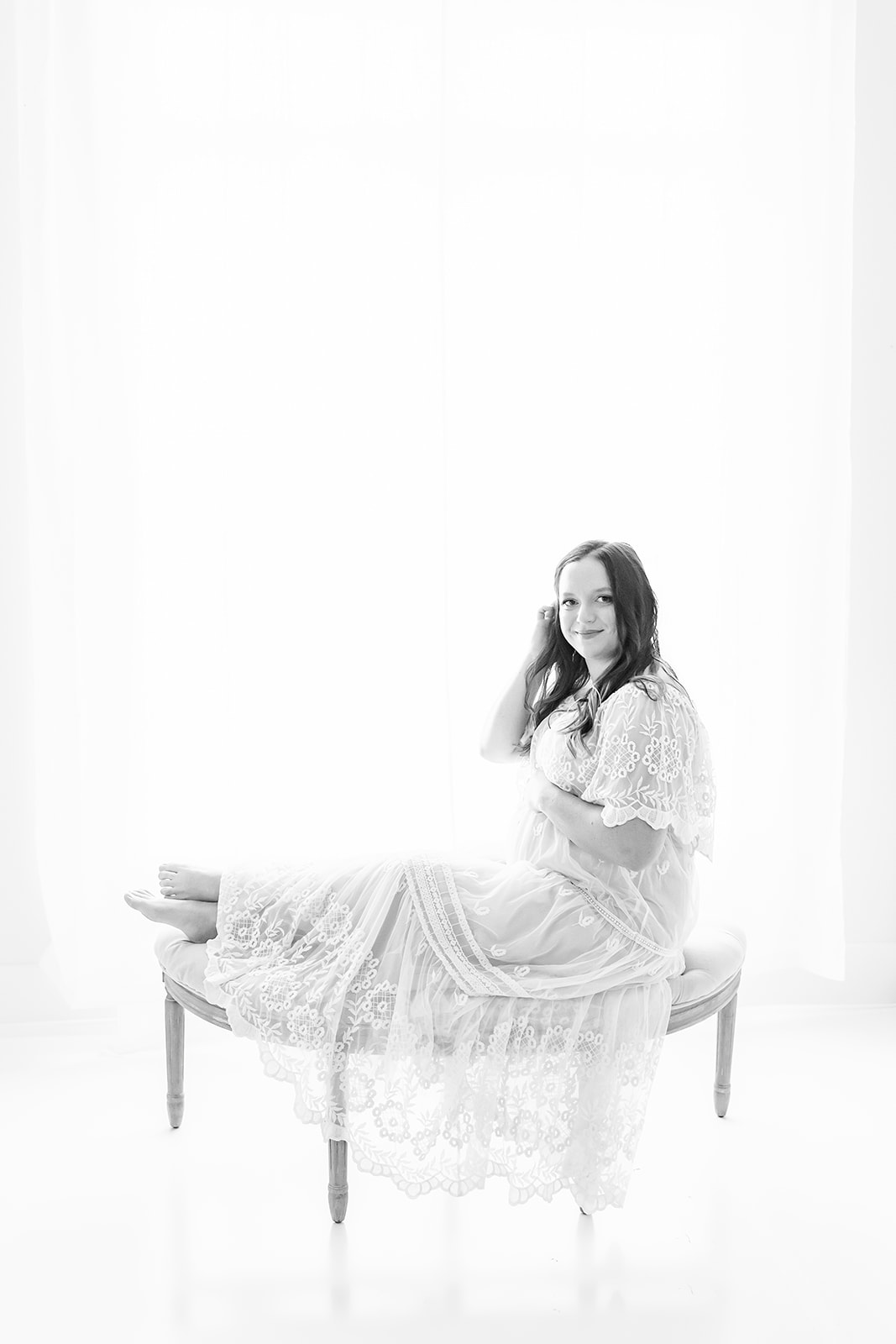 A mother to be sits on a curved bench in a lace dress in front of a window in a studio Pittsburgh midwife