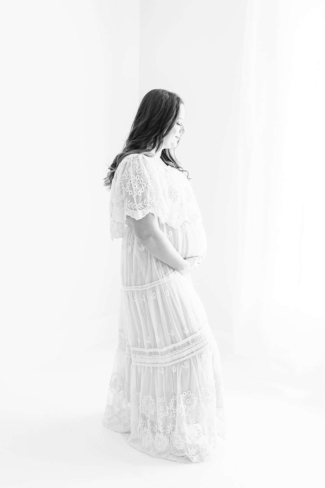 A mom to be stands in a studio by a window wearing a white lace maternity dress Pittsburgh midwife