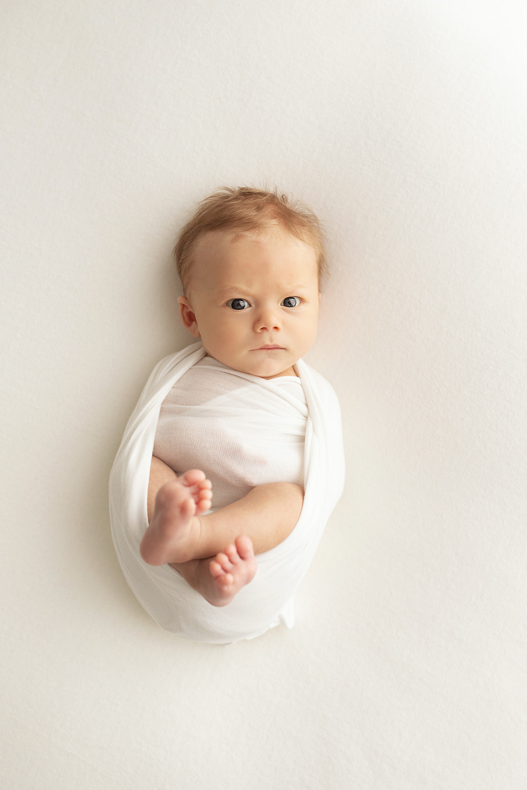 A newborn baby lays on a white bed in a white swaddle with eyes open Pittsburgh Nannies