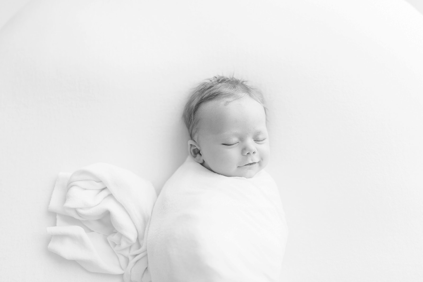 A newborn baby sleeps with a smile in a white swaddle