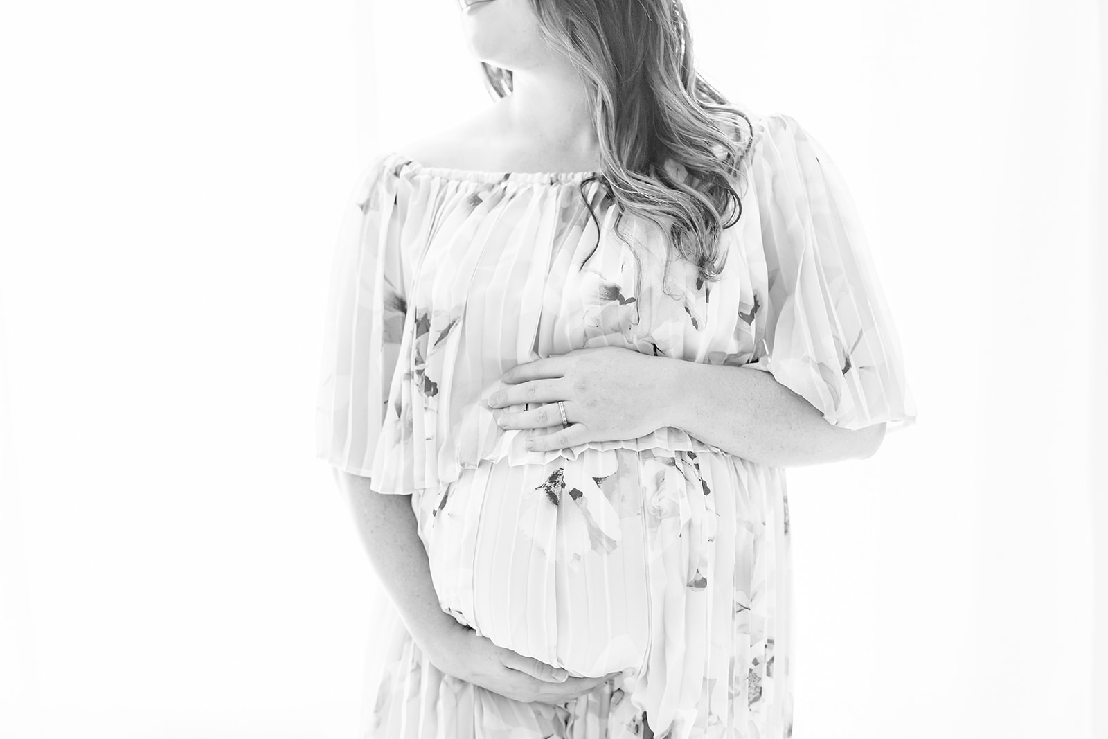 A mother to be in a floral pattern maternity dress stands in a studio holding her bump BHS Women’s Care Associates