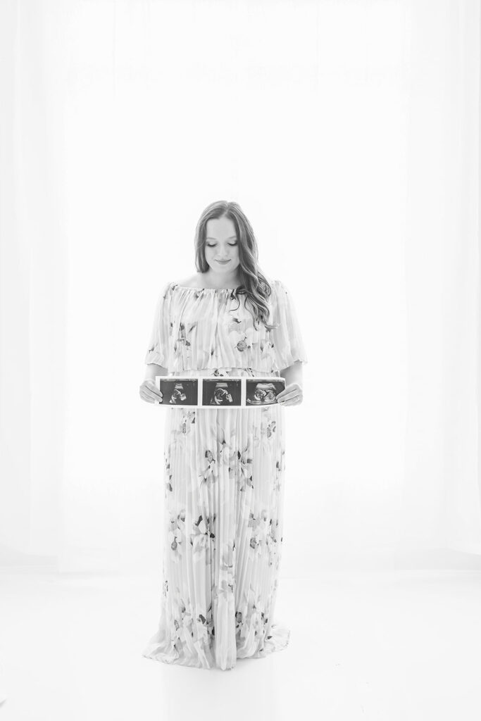 A mother to be in a floor-length maternity dress stands in a studio holding her sonogram BHS Women’s Care Associates