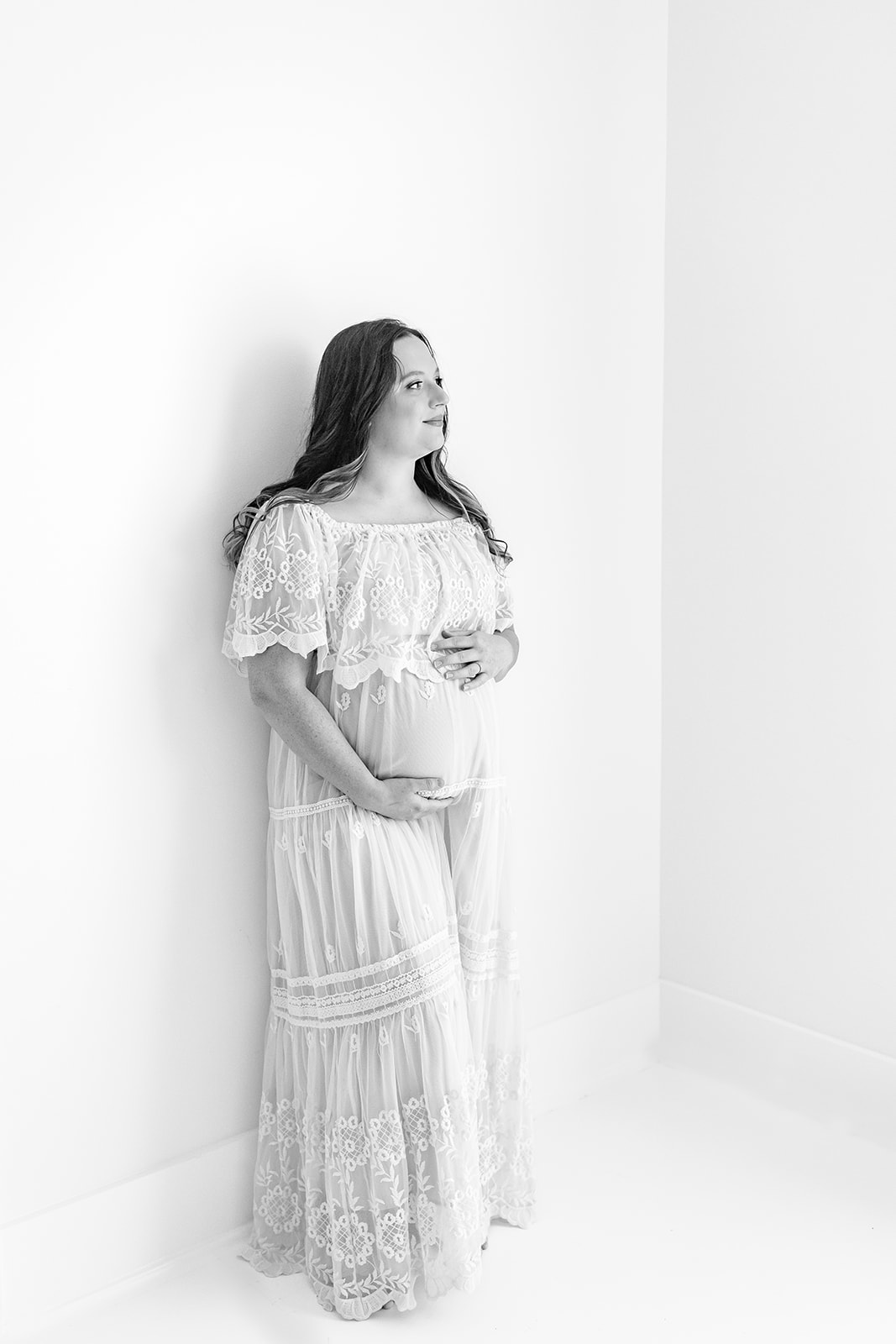 A mother to be leans against a wall in a studio while holding her bump in a white maternity gown