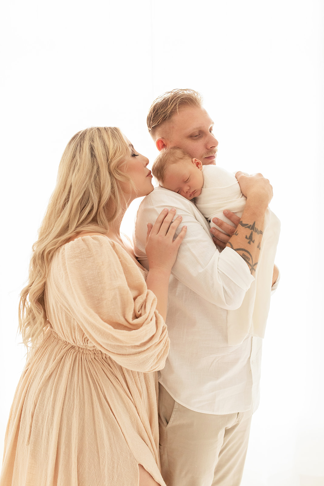 A newborn baby sleeps on dad's shoulder while mom leans in to kiss the baby while standing in a studio