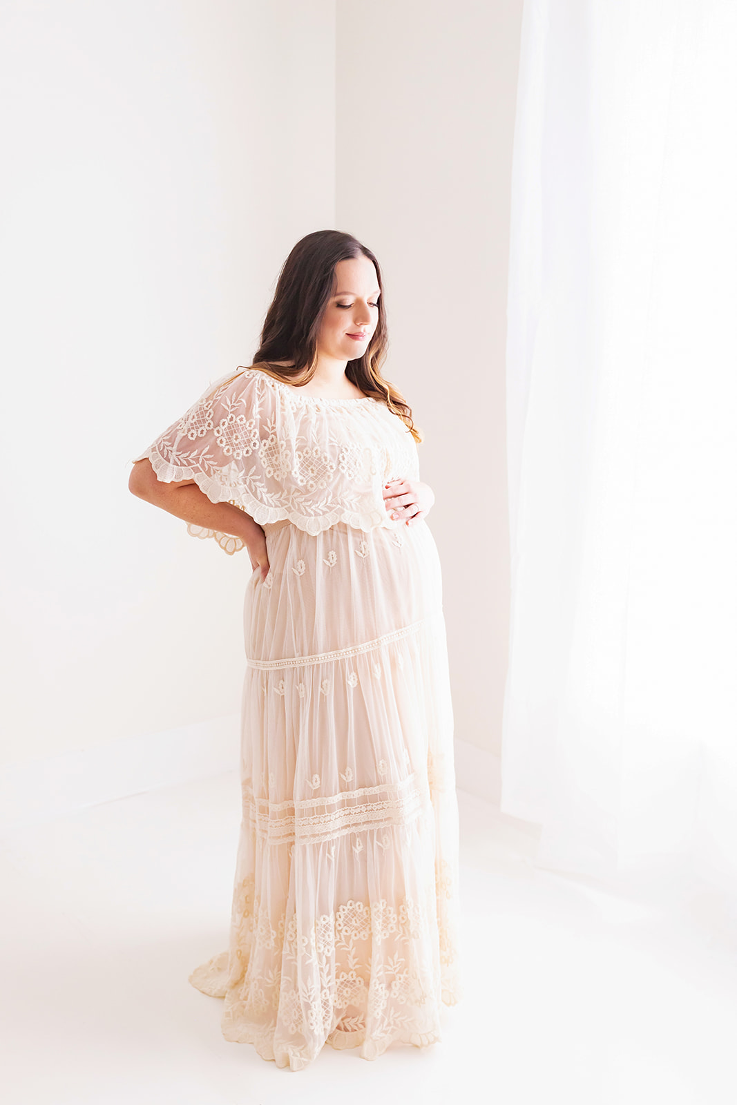 A mother to be rests a hand on her bump while looking down at it while standing in a studio