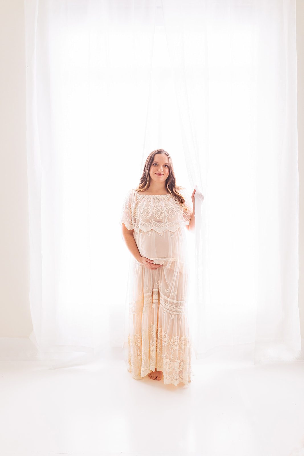 A mother to be in a beige lace maternity gown stands in front of a large window in a studio Pittsburgh Prenatal Massage