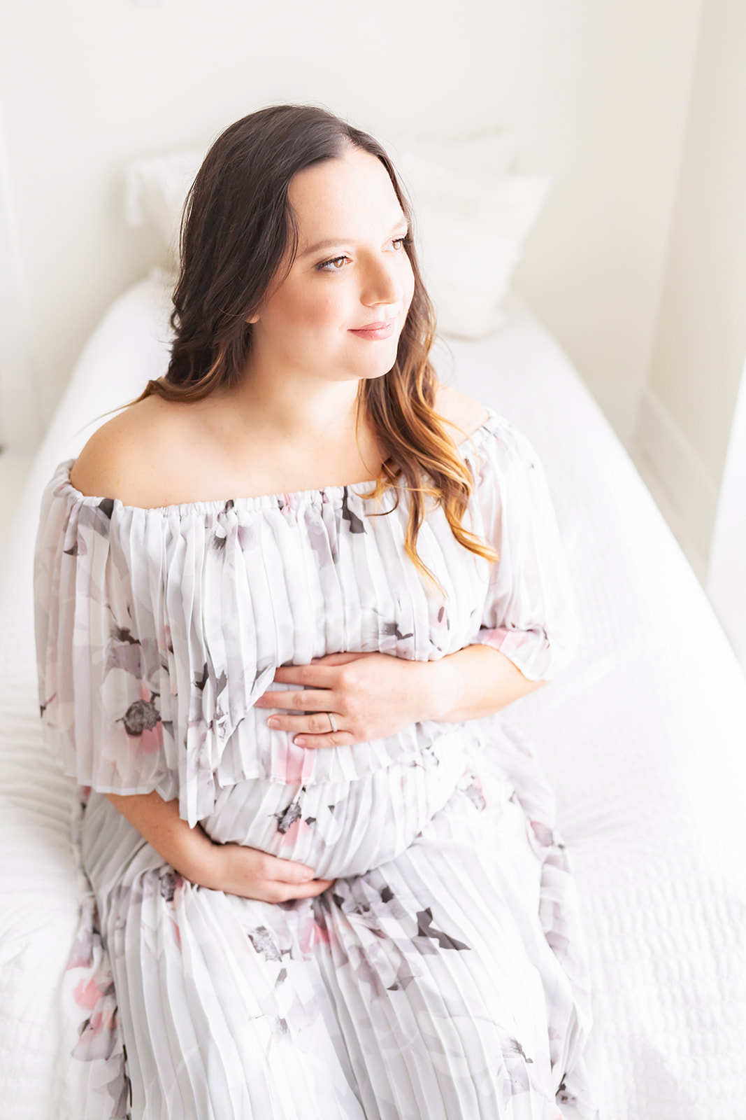 A mom to be sits on the edge of a bed holding her bump gazing out a window