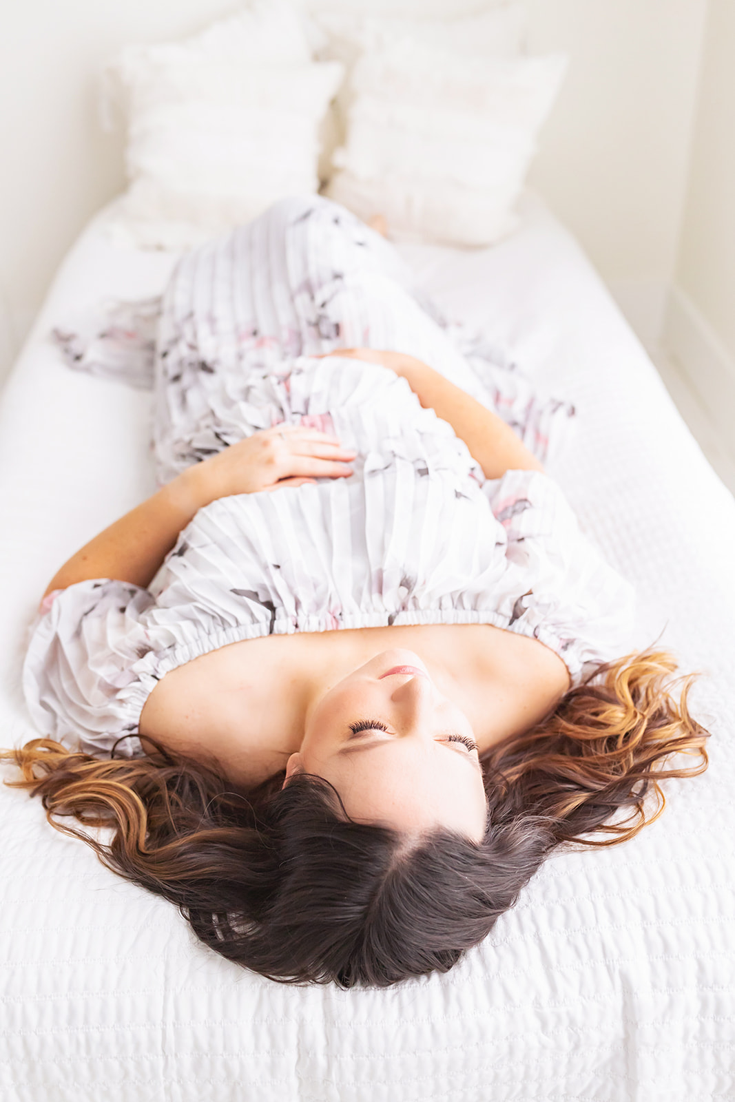 A mom to be lays across a bed holding her bump on her back while wearing a white maternity gown Pittsburgh baby shower venues