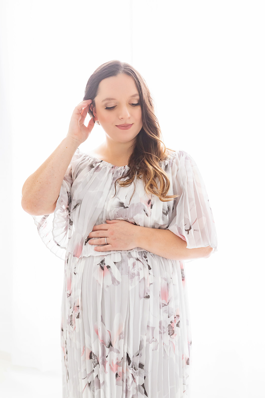 A mom to be in a floral maternity gown runs a finger through her hair while standing in front of a large window