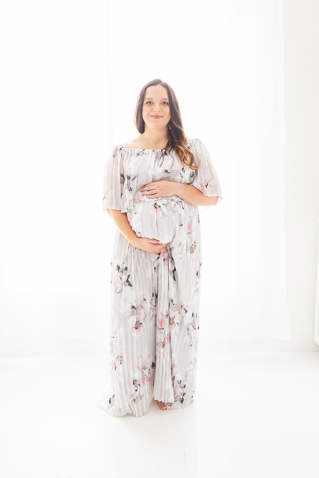 A mom to be stands in a studio in front of a large window holding her bump in a floor length maternity gown Pittsburgh OBGYN