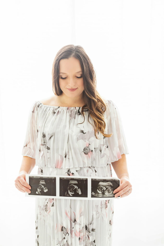 A mom to be stands in a studio looking down at her sonograms in her hands held out in front of her Pittsburgh OBGYN