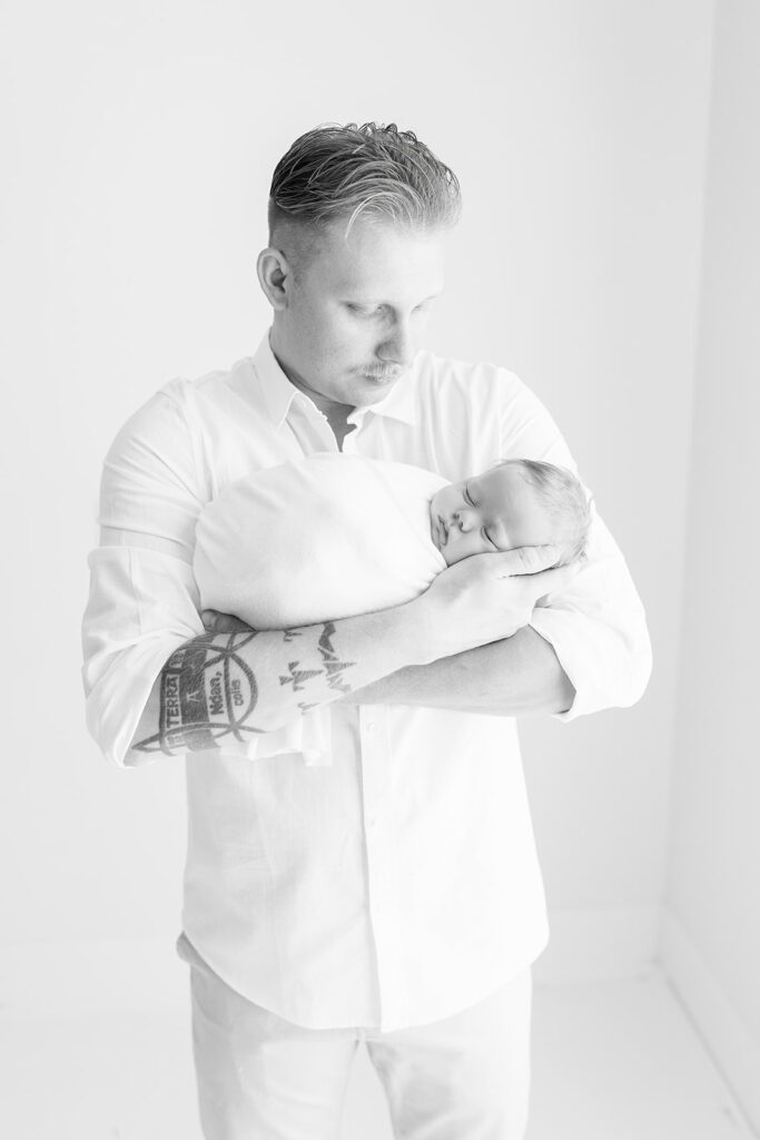 A new father gazes down at his newborn baby in his arms while standing by a window Pittsburgh Pediatricians