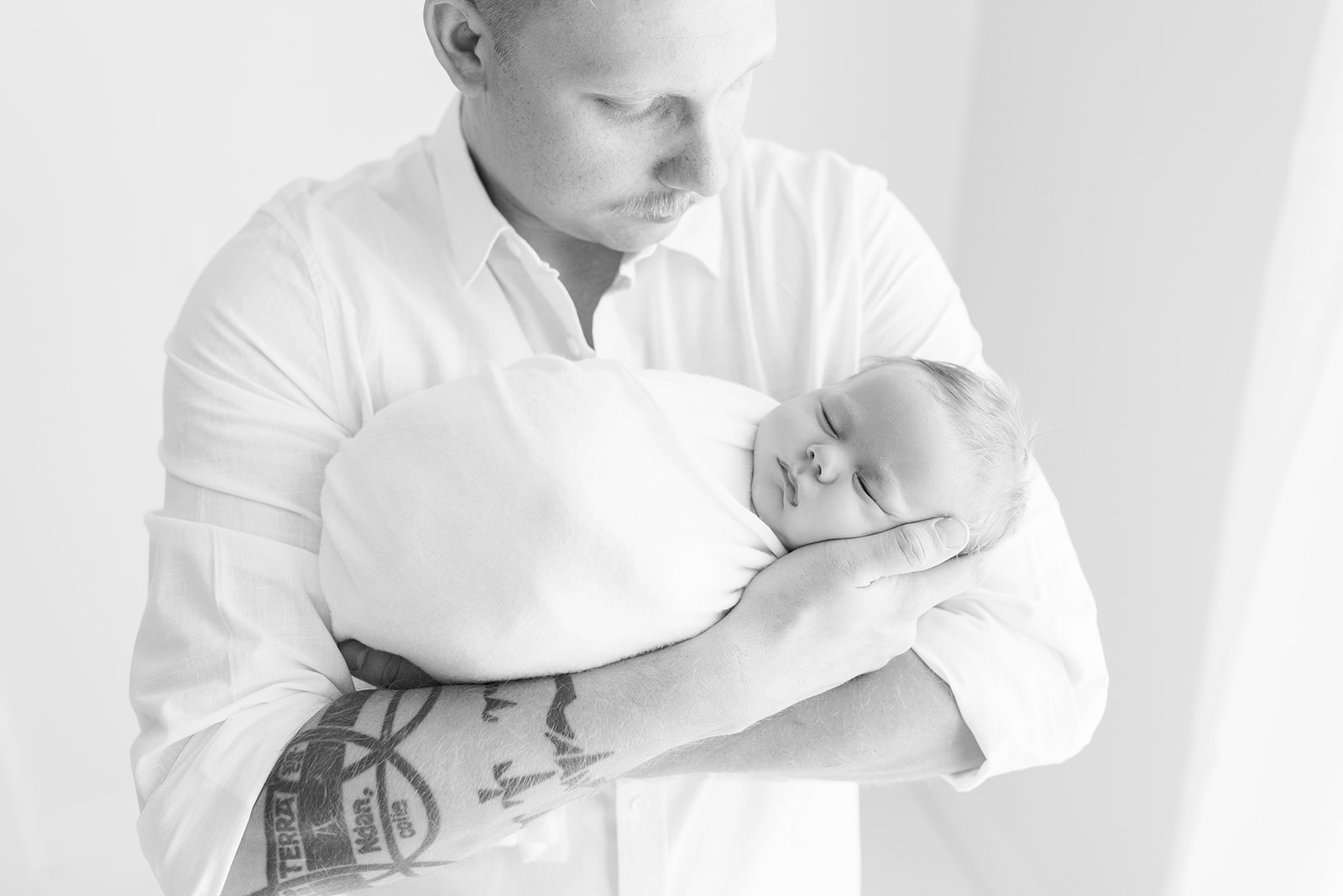 A father in a white shirt looks down at his sleeping newborn baby in his arms while standing in a studio Pittsburgh Pediatricians