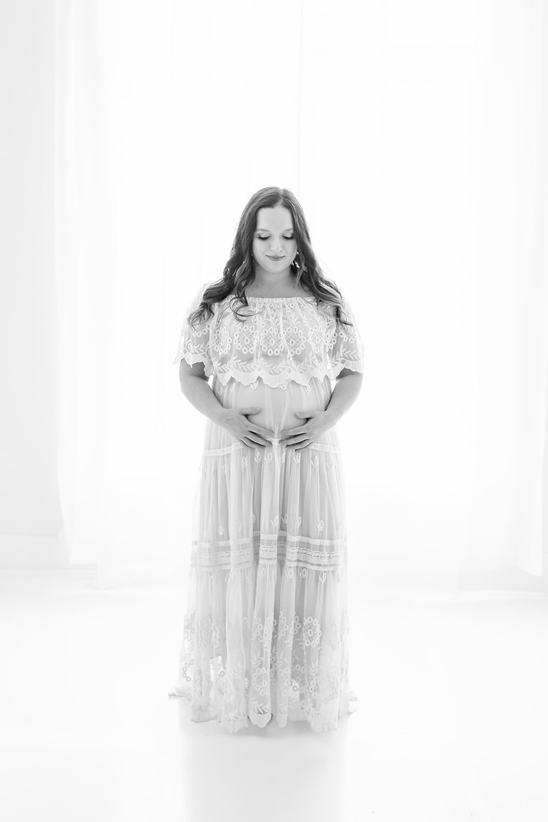 A mom to be smiles down at her bump while resting her hands on it in a studio