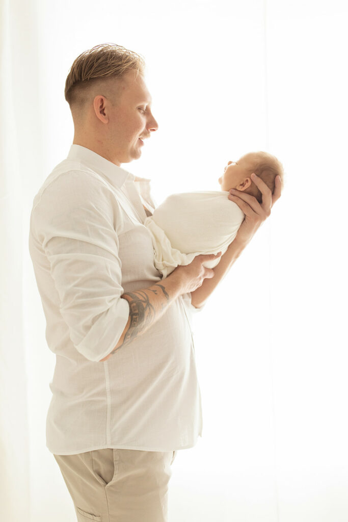 A new father in a white shirt stands in a window looking down at his newborn baby in his hands Little Gym Pittsburgh