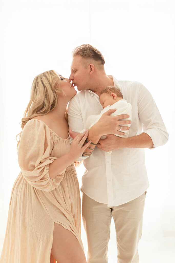 New parents kiss while standing in a studio holding their sleeping newborn baby