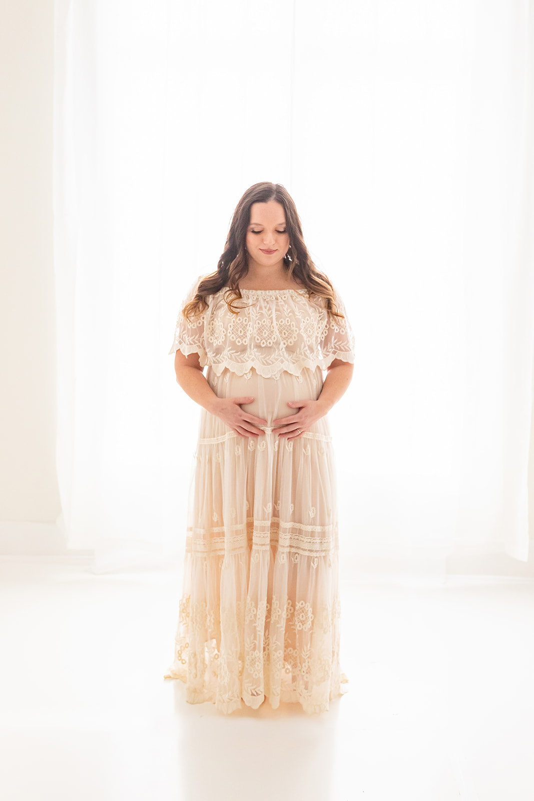 A mom to be smiles down to her bump while standing in a studio in front of a large window