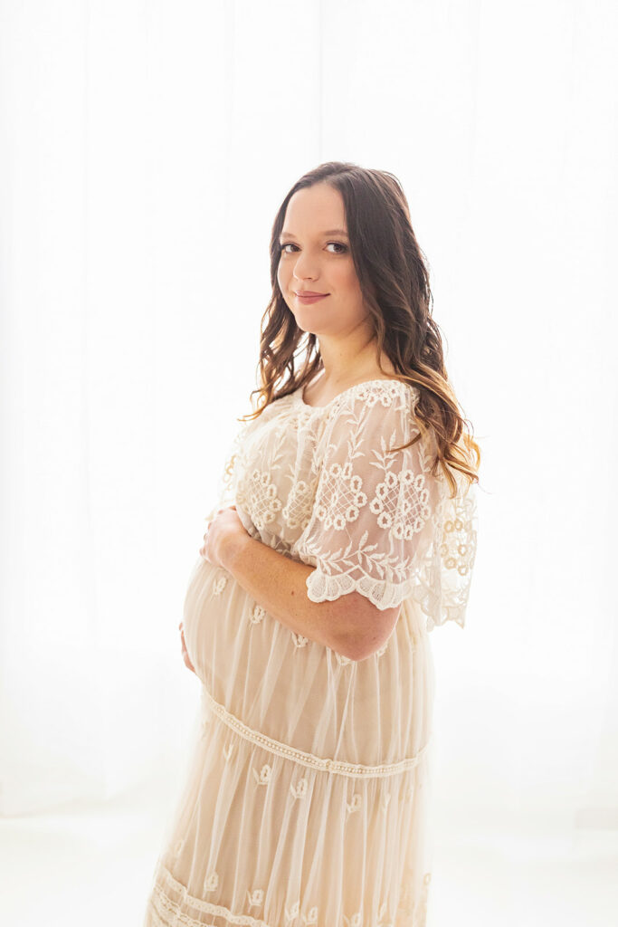 A mom to be stands in a studio in a lace maternity gown holding her bump Prenatal chiropractor Pittsburgh