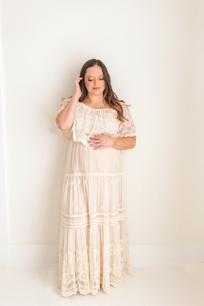 A mom to be in a white lace maternity gown runs a finger through her hair while standing in front of a white wall
