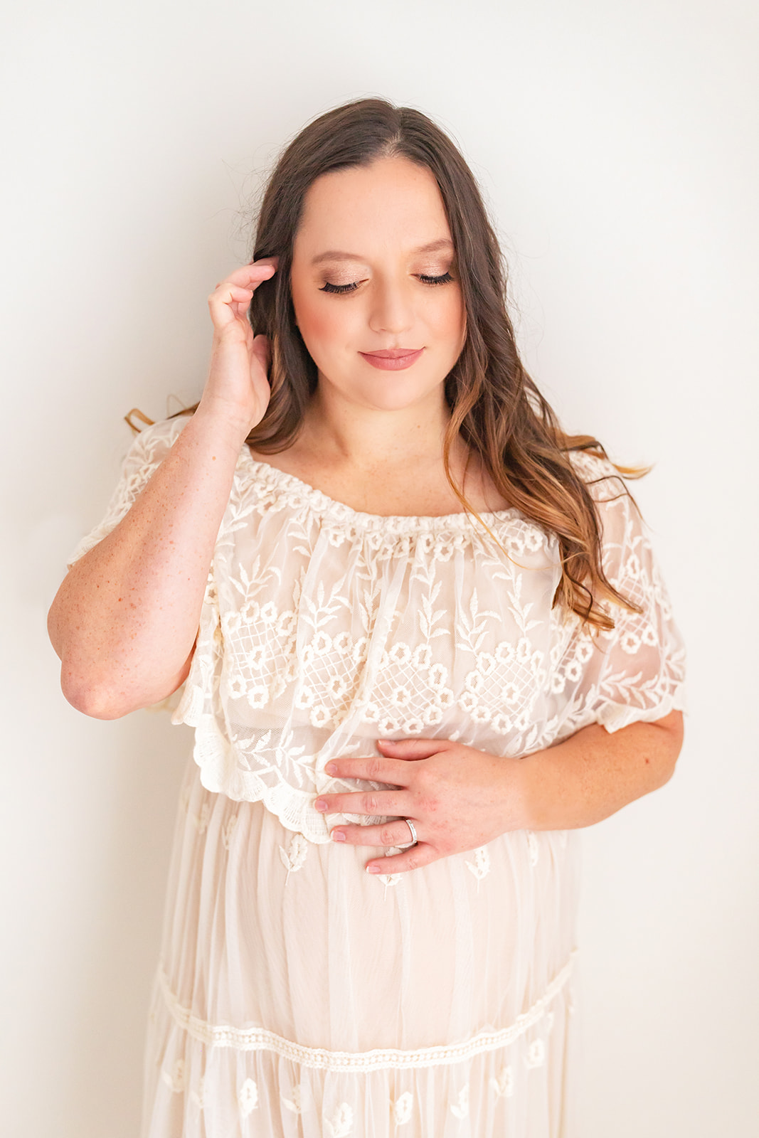 A mom to be in a white lace maternity gown runs a finger through her hair while standing in front of a white wall babymoon ideas near Pittsburgh