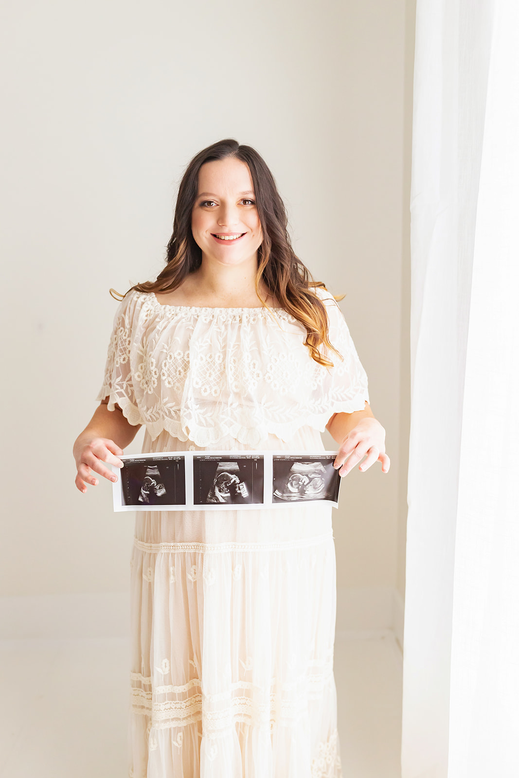 A mother to be in a white lace dress stands in a window smiling while holding her sonograms babymoon ideas near Pittsburgh