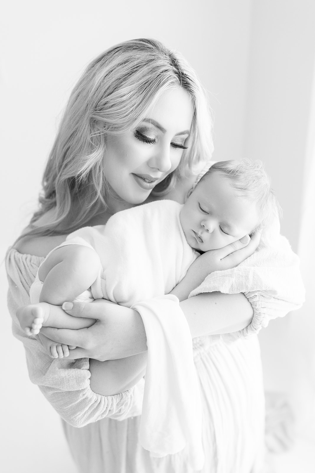 A new mother in a white dress stands in a window while cradling her sleeping newborn baby in her arms