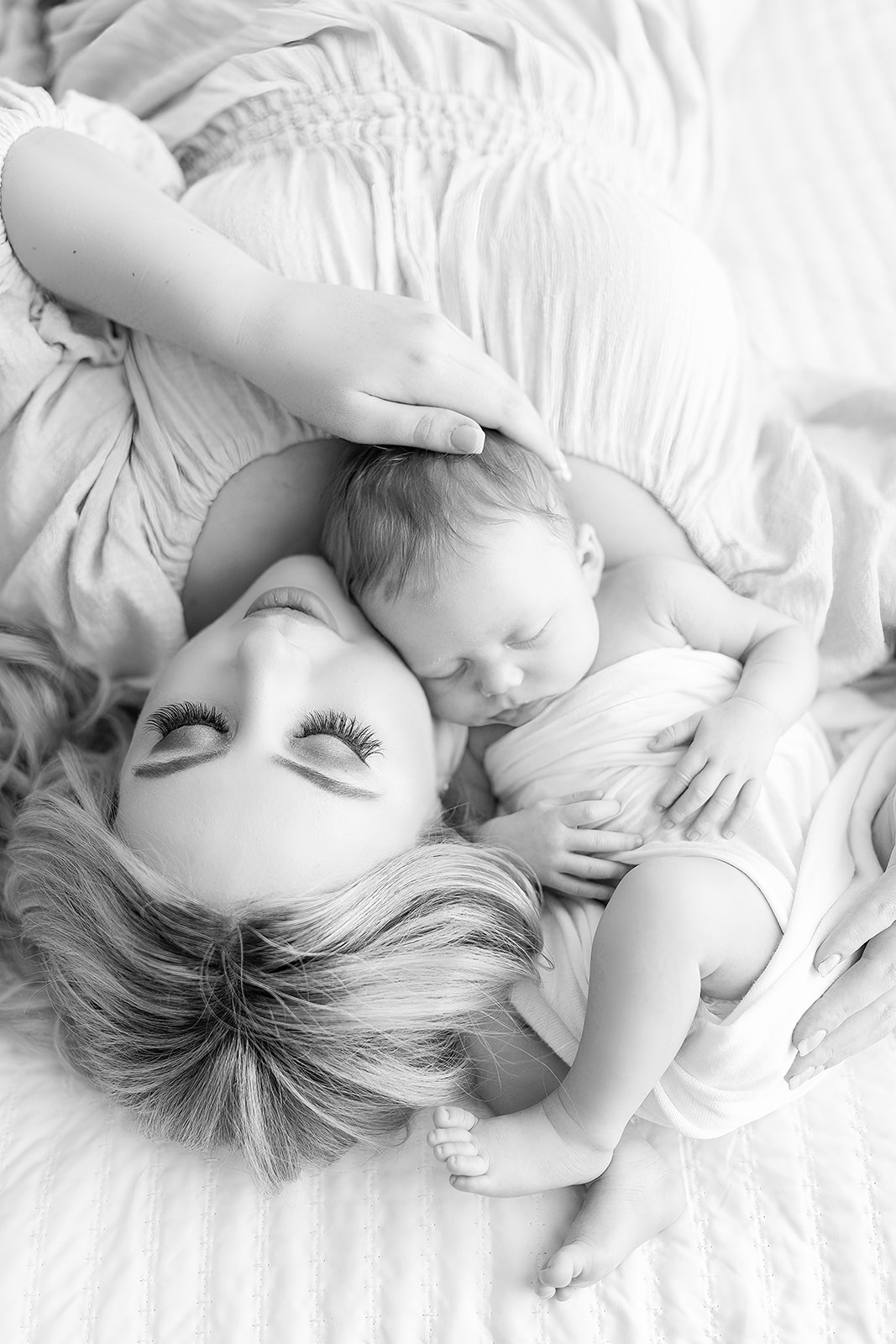 A new mom lays on a white bed with her newborn baby wrapped in a white blanket snuggling on her shoulder playgrounds in Pittsburgh