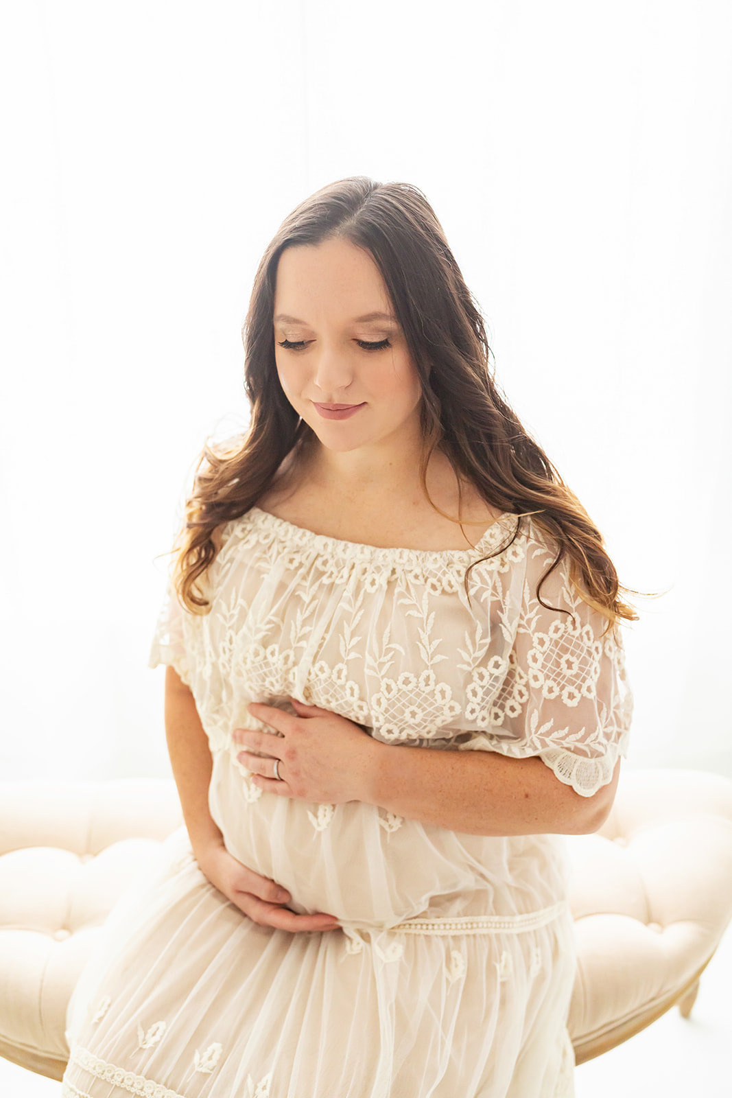 A mom to be sits on the edge of a bench in a studio holding her bump in a beige lace maternity gown