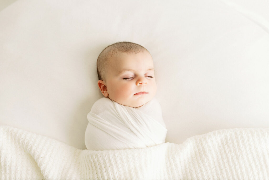 A newborn baby sleeps by a window in a studio wrapped tightly in a white swaddle thanks to baby sleep consultant pittsburgh