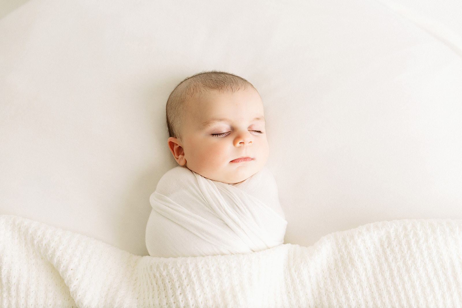 A newborn baby sleeps by a window in a studio wrapped tightly in a white swaddle thanks to baby sleep consultant pittsburgh