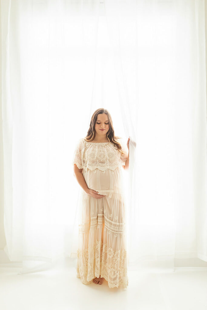 A pregnant woman stands in the curtains of a window in a beige maternity gown while holding her bump with help from Birthing Classes Pittsburgh