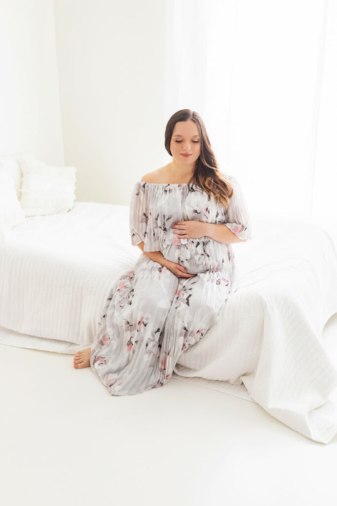 A mom to be in a floral dress sits on the edge of a bed in a studio by a window after getting help from Bloom Birth Concierge