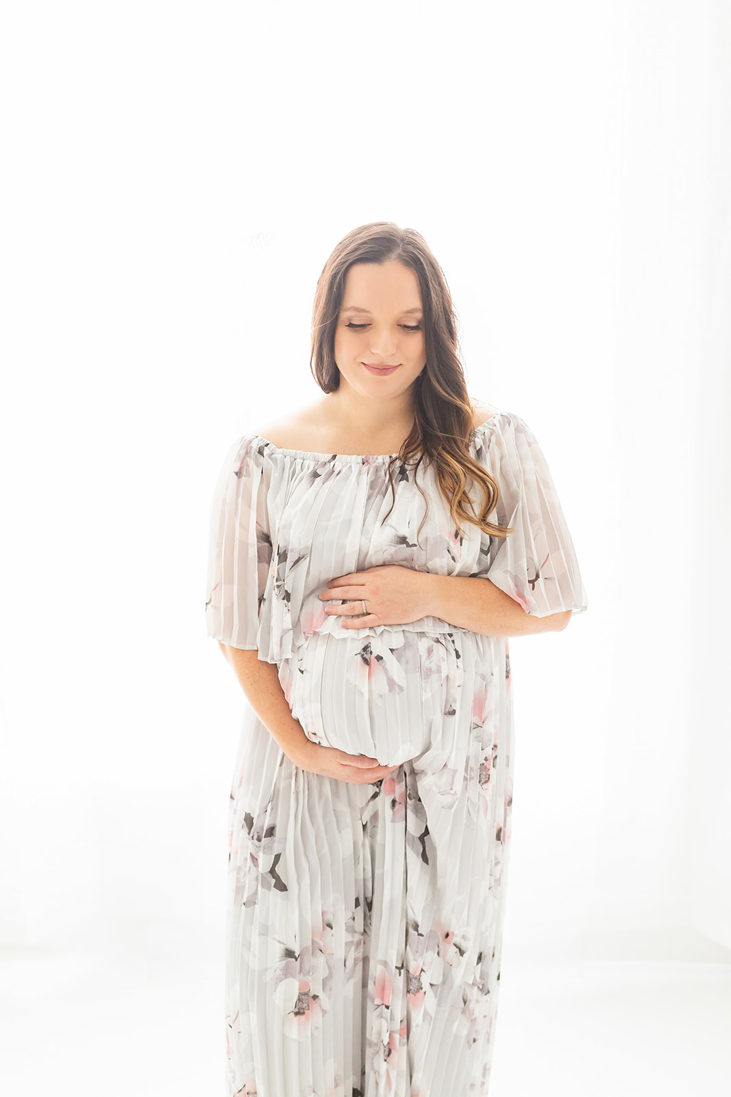 A mom to be in a floral dress smiles down to her bump while standing in a window in a studio after visiting Bloom Birth Concierge