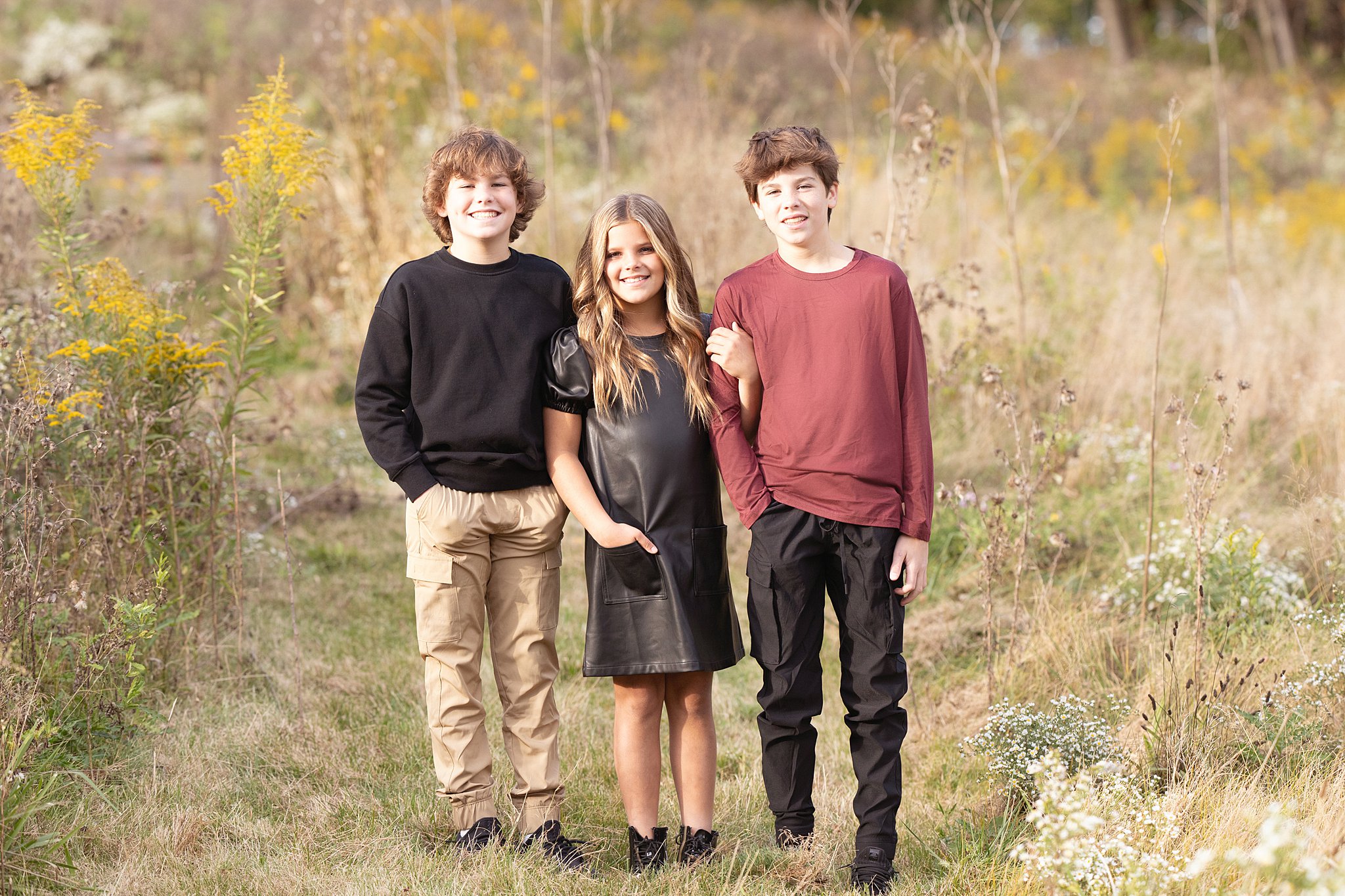 Three siblings stand in a trail in a grassy field smiling before attending Childrens Museum of Pittsburgh