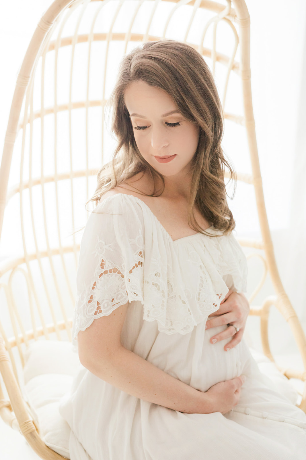 A mom to be in a white maternity gown sits on a wicker chair in a studio smiling down her shoulder thanks to IVF Pittsburgh
