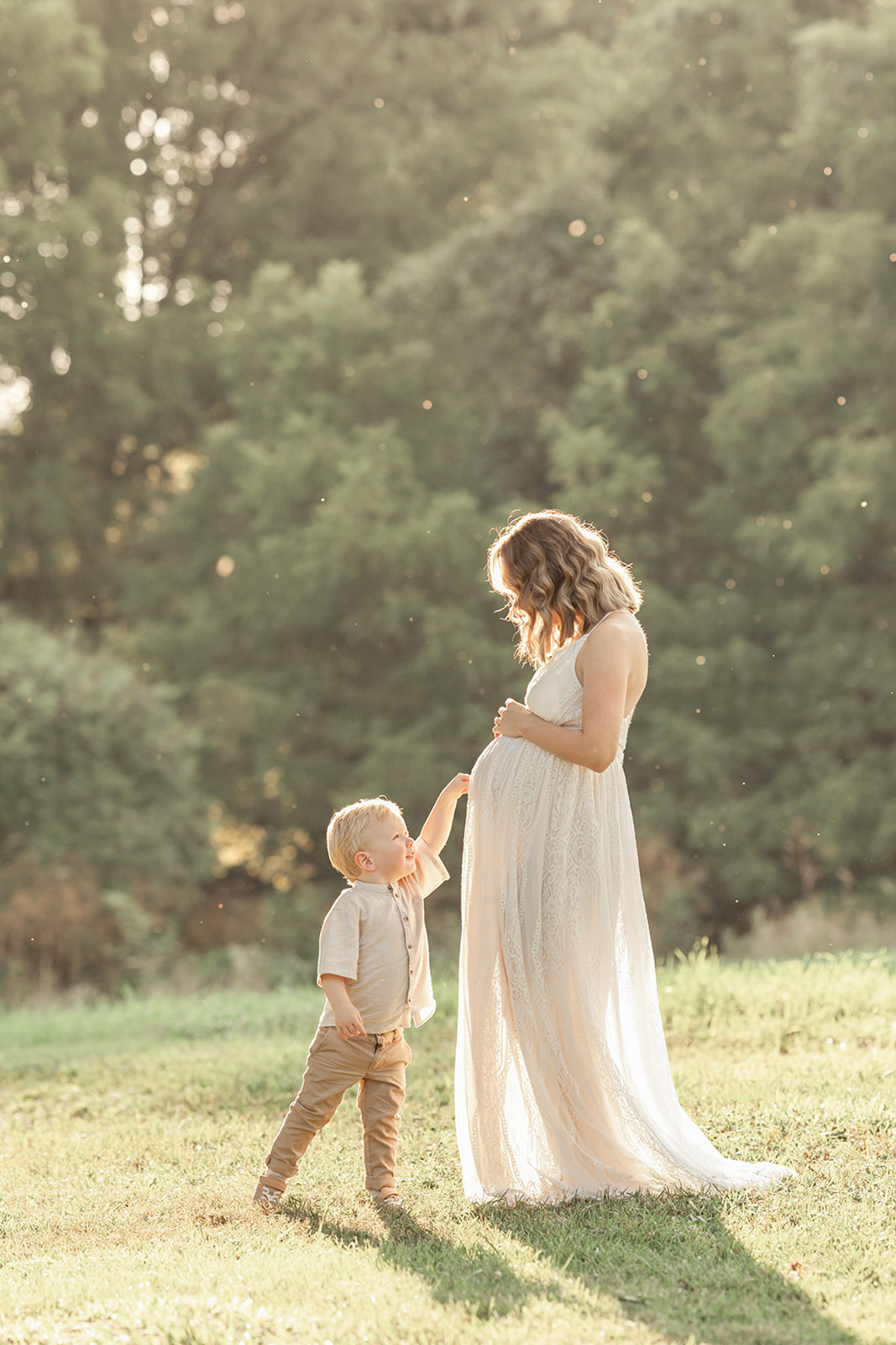 A pregnant mother in a white lace maternity dress stands playing in a park with her toddler son before visiting Lula boutique pittsburgh