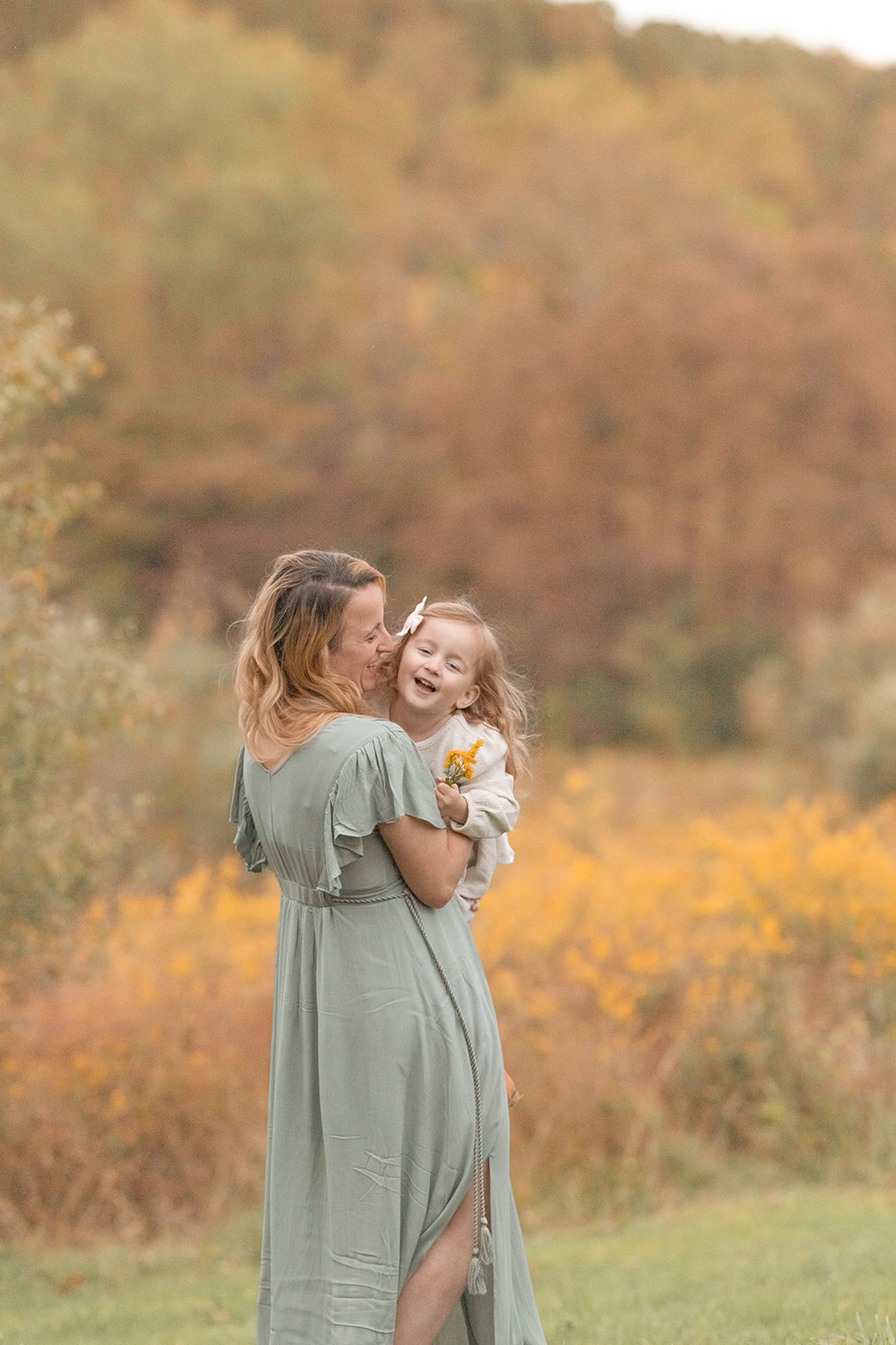 A mom in a green dress tickles and plays with her young daughter in a field of wildflowers after visiting a Montessori preschool Pittsburgh